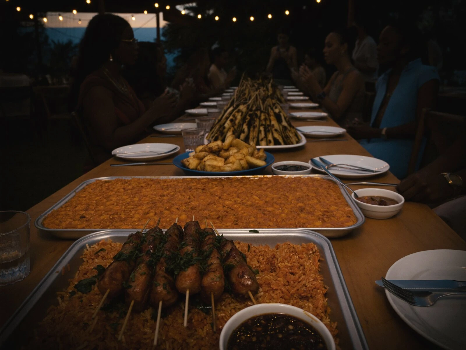 Long communal table set for an outdoor event at dusk, with jollof rice, suya skewers, fried plantain and pepper sauce, guests seated together under warm string lights