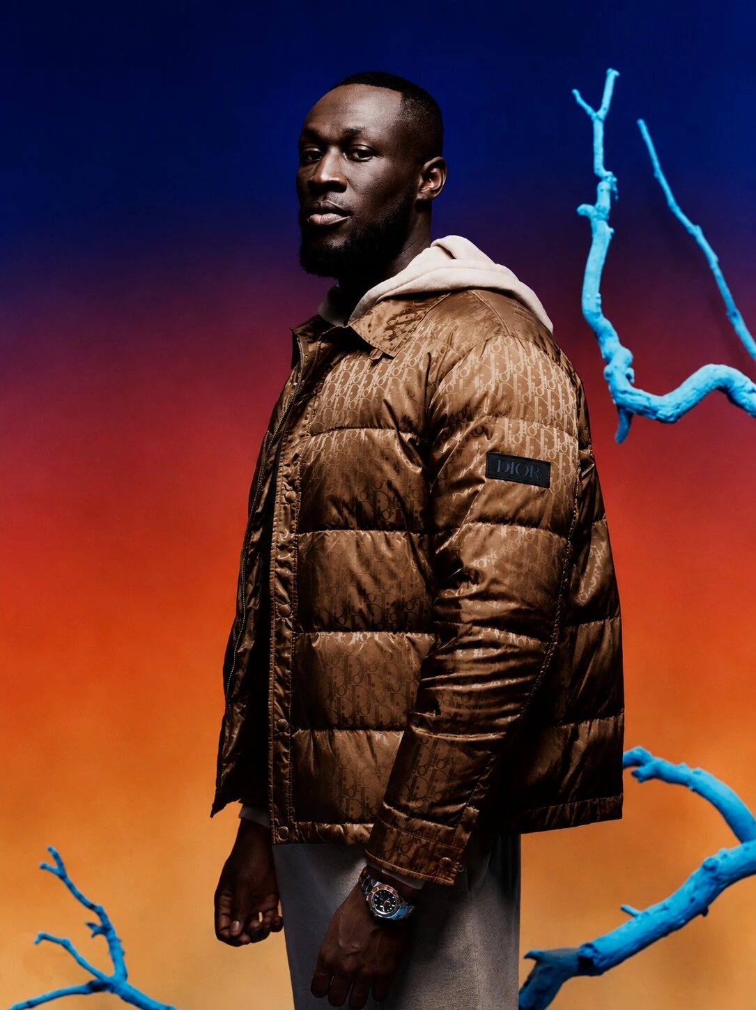 Stormzy in a bronze puffer jacket against a blue to orange backdrop with blue branches