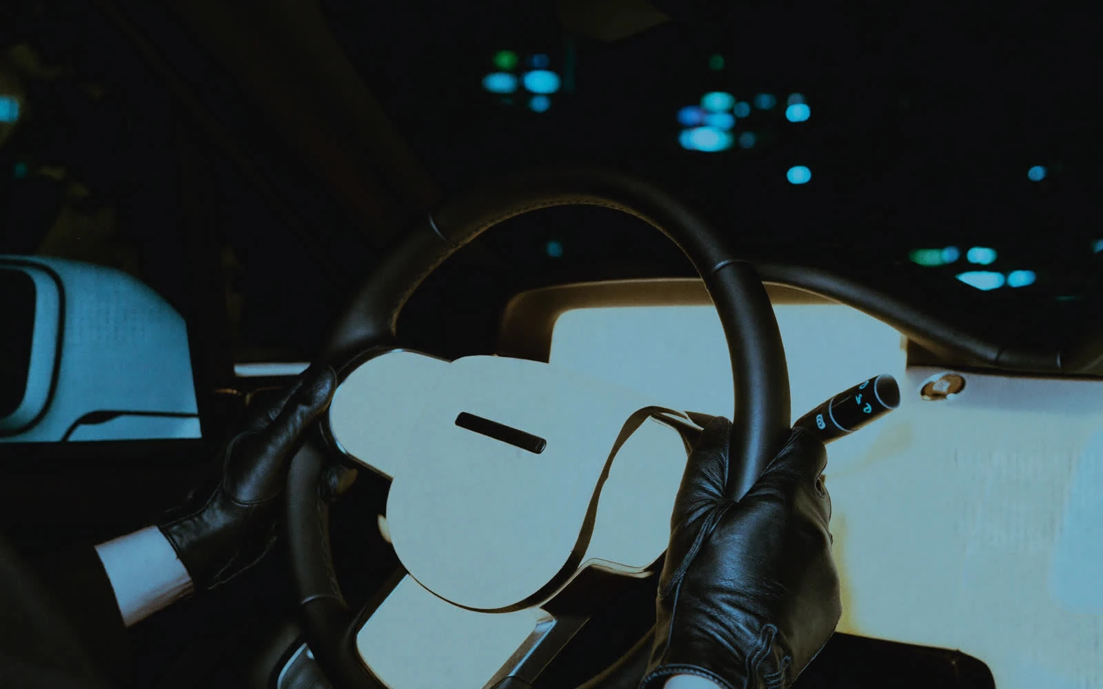 Gloved chauffeur hands resting on the leather steering wheel at night, dashboard glow and blurred city lights beyond the windscreen.