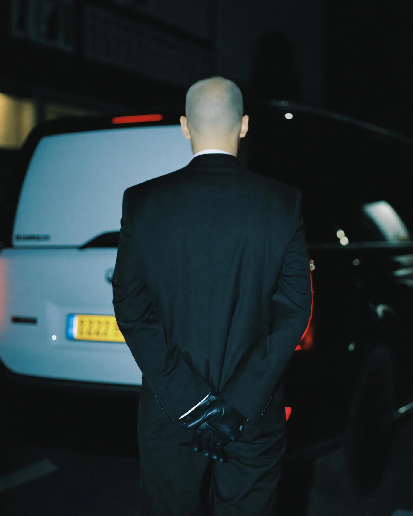 Chauffeur’s gloved hands resting on the wheel, a flight-tracking display glowing cool steel-blue on the dashboard at dusk